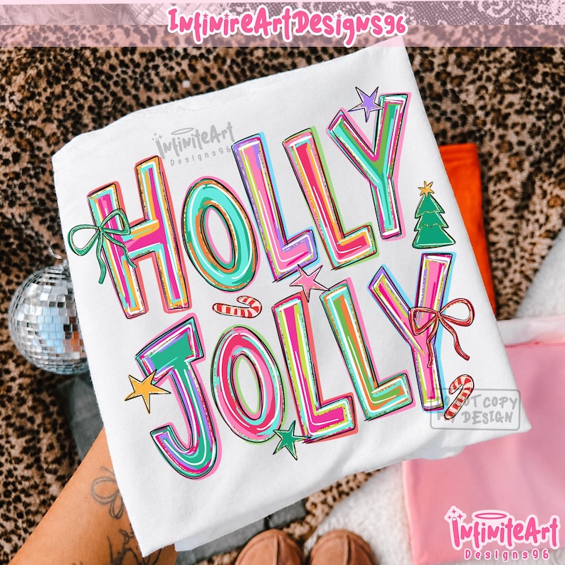 May include: A white t-shirt featuring the text HOLLY JOLLY in large, colorful, outlined letters. The design includes a green Christmas tree, a small candy cane, a yellow star, and two bows in green and red, all printed on the front of the shirt.