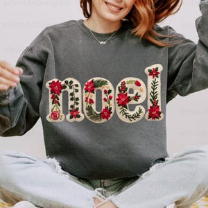 May include: A dark gray sweatshirt with the word "noel" embroidered across the front. The letters are decorated with red poinsettias, green leaves, and white accents, creating a festive holiday design.