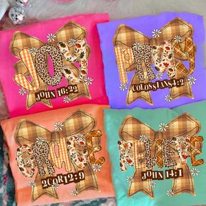 Op de afbeelding: Vier kleurrijke t-shirts met inspirerende boodschappen. Elk shirt heeft een groot strikdesign met de woorden "JOY", "PRAY", "GRACE" en "BELIEVE" in een mix van patronen en lettertypen. Bijbelteksten staan onder elk woord gedrukt.