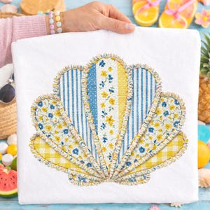 May include: White beach towel featuring a seashell design. The seashell is composed of various patterned fabrics, including blue stripes, yellow gingham, and floral prints. The edges are trimmed with a ruffled fabric. The towel is held by a person.