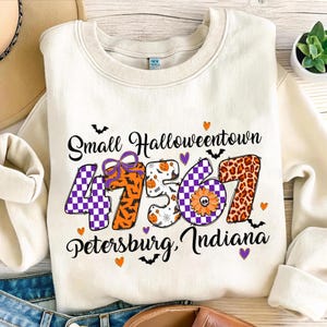 Könnte beinhalten: Cremefarbenes Sweatshirt mit dem Text "Small Halloweentown 47567 Petersburg, Indiana". Die Zahlen sind mit Mustern wie lila Schachbrett, orange Fledermäusen und Leopardenmuster verziert. Halloween-Kleidung.