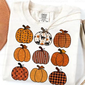 May include: White Comfort Colors t-shirt featuring nine different pumpkin designs. The pumpkins have various patterns, including bats, cats, spiderwebs, and plaid. The shirt is a small size.