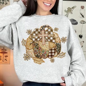 Puede incluir: Una sudadera gris claro con un dise&ntilde;o de calabaza de retazos. La calabaza tiene un lazo a cuadros, una cruz de madera y detalles florales y de hojas. El dise&ntilde;o est&aacute; centrado en la parte delantera de la sudadera.
