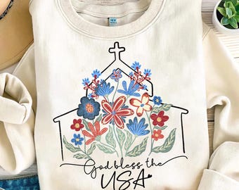 May include: Cream-colored sweatshirt featuring a black outline of a church with a cross on top, filled with red, blue, and white floral designs. The words "God bless the USA" are written in a script font below the church illustration.