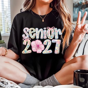 Puede incluir: Camiseta negra con la frase "Senior 2021" en un dise&ntilde;o floral. La palabra "Senior" es de color azul claro con un estampado floral, y los n&uacute;meros "2021" son de un estilo similar. Una gran flor rosa est&aacute; entre los n&uacute;meros.