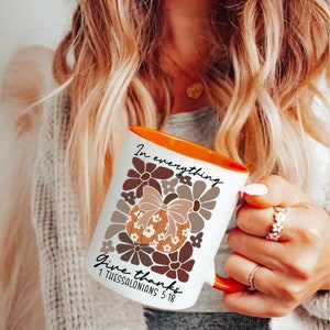 May include: White ceramic mug with an orange rim. The mug features a brown and orange floral design with a pumpkin in the center. The text "In everything give thanks 1 Thessalonians 5:18" is printed on the mug.