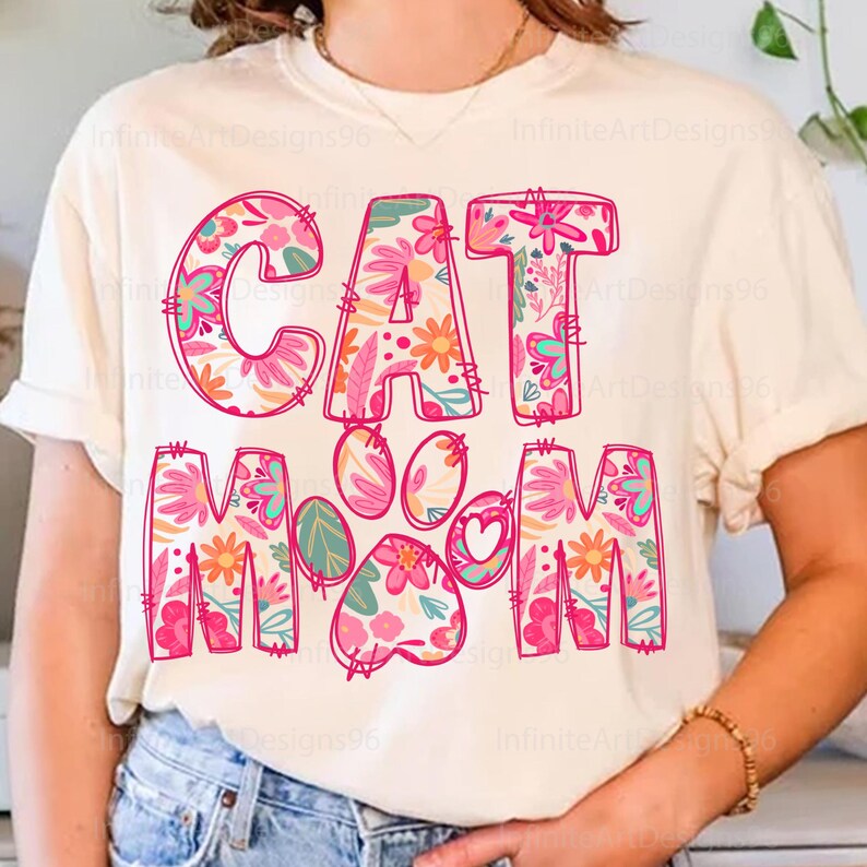 May include: A white t-shirt with the text "CAT MOM" in pink outlined letters. The letters are filled with a floral pattern in pink, orange, and green. A pink paw print with a heart in the center is between the words "CAT" and "MOM".