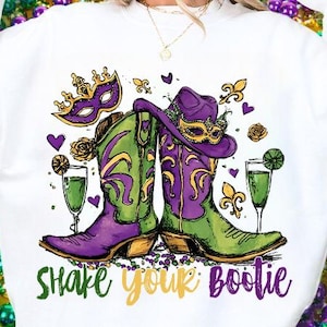 Puede incluir: Sudadera blanca con un colorido diseño de Mardi Gras. Presenta botas de vaquero verdes y moradas, un sombrero de vaquero morado, una máscara y el texto "Shake Your Bootie". Incluye copas de champán y elementos decorativos.