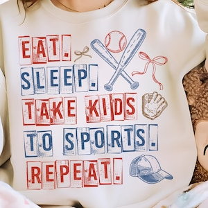 May include: Cream-colored sweatshirt with a baseball theme. The front features the text "EAT. SLEEP. TAKE KIDS TO SPORTS. REPEAT." in red and blue block letters, along with baseball bats, a ball, a glove, and a cap.