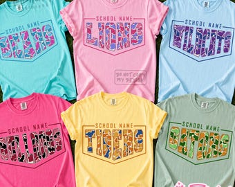 Könnte beinhalten: Sechs kurzärmelige T-Shirts mit Rundhalsausschnitt in Türkis, Pink, Hellblau, leuchtendem Pink, Gelb und Salbeigrün. Jedes Shirt zeigt eine Grafik mit dem Text SCHOOL NAME über den Teamnamen BEARS, LIONS, WILDCATS, BULLDOGS, TIGERS und GATORS.