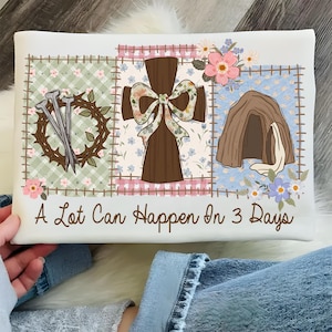May include: White rectangular cushion with a patchwork design featuring a cross, nails, a crown of thorns, and a tomb. The cushion has the text "A Lot Can Happen In 3 Days" in a script font.