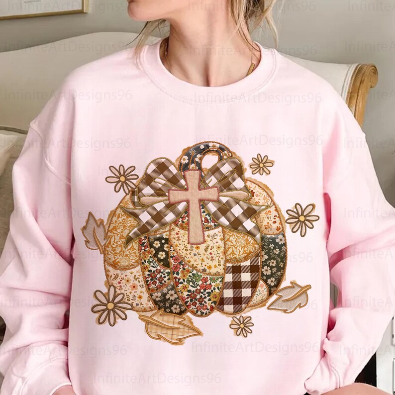 Puede incluir: Una sudadera rosa claro con un dise&ntilde;o de calabaza de retazos. La calabaza tiene una cruz y un lazo a cuadros. Acentos florales y de hojas rodean la calabaza. El texto "Infinite Art Designs96" es visible.