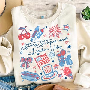 May include: Cream-colored sweatshirt with a patriotic design featuring cherries, fireworks, an American flag, and the text "Stars Stripes and Freedom Vibes." The design incorporates red and blue elements.
