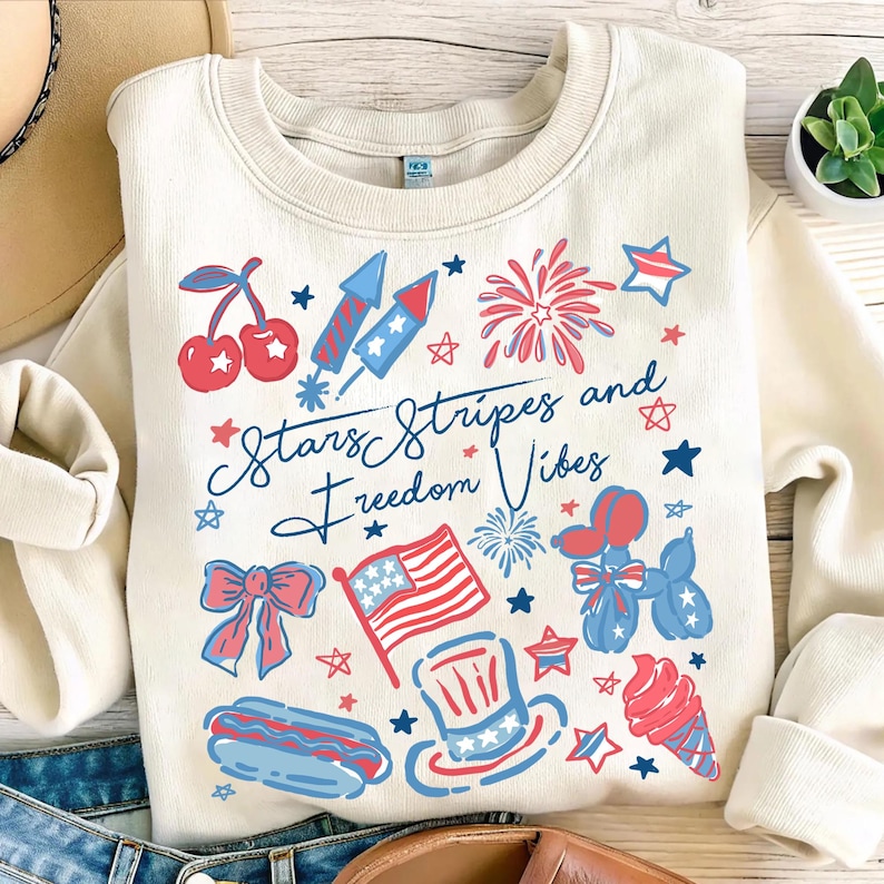 May include: Cream-colored sweatshirt with a patriotic design featuring cherries, fireworks, an American flag, and the text "Stars Stripes and Freedom Vibes." The design incorporates red and blue elements.