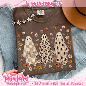 May include: A brown t-shirt featuring a graphic of three ghosts standing among colorful flowers. The first ghost has a checkered pattern, the second has a leopard print, and the third has black polka dots. The shirt has a crew neckline.