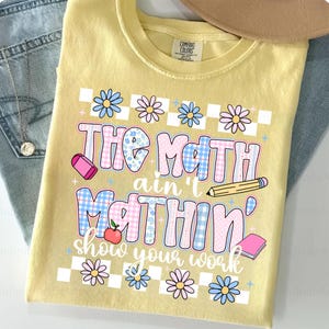 May include: Yellow Comfort Colors t-shirt with the phrase "The Math Ain't Mathin' Show Your Work" in a playful font. The design includes daisies, an apple, a pencil, an eraser, and a book. The shirt is paired with jeans and a necklace.