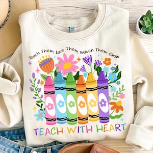 Może przedstawiać: Kremowa bluza z kolorowym wzorem kredek i kwiatów. Grafika zawiera tekst "Teach Them, Love Them, Watch Them Grow" i "Teach With Heart". Kredki są różowe, niebieskie, zielone, pomarańczowe, żółte i fioletowe.