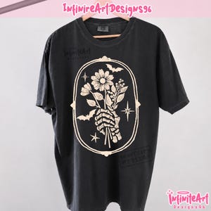 Pode incluir: Uma t-shirt preta de manga curta com um gráfico em cor creme de uma mão esquelética segurando um ramo de flores dentro de uma moldura oval. O design inclui pequenos morcegos e estrelas de quatro pontas. O texto InfiniteArtDesigns96 é visível.