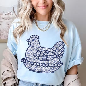 May include: Light blue t-shirt featuring a whimsical hen design. The hen is adorned with a blue floral pattern and sits on a decorative base. The shirt has short sleeves and a relaxed fit, perfect for casual wear. The design is centered on the front of the shirt.