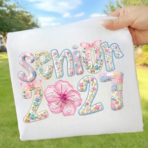 Puede incluir: Camiseta blanca con la palabra "SENIOR" y el n&uacute;mero "2021" bordados con un dise&ntilde;o floral y patchwork. Una gran flor rosa tambi&eacute;n est&aacute; bordada en la camiseta. La camiseta est&aacute; sujeta por una mano.