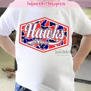 Op de afbeelding: Een wit T-shirt met korte mouwen en een centrale zeshoekige afbeelding met een rood, wit en blauw camouflagepatroon. De afbeelding toont het woord Hawks in een wit scriptlettertype, met de tekst Hardin Jefferson eronder gedrukt.