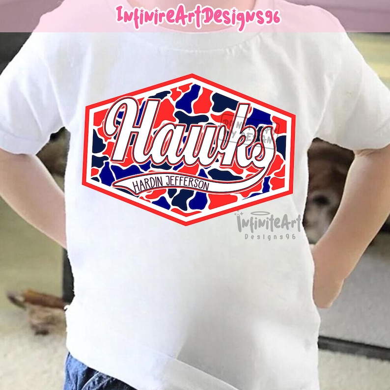 Op de afbeelding: Een wit T-shirt met korte mouwen en een centrale zeshoekige afbeelding met een rood, wit en blauw camouflagepatroon. De afbeelding toont het woord Hawks in een wit scriptlettertype, met de tekst Hardin Jefferson eronder gedrukt.