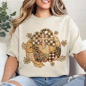 Puede incluir: Camiseta color crema con un dise&ntilde;o de calabaza de retazos, una cruz, un lazo y detalles florales y de hojas. La calabaza est&aacute; compuesta por varias telas estampadas. El texto "Infinite Art Designs96" es visible.