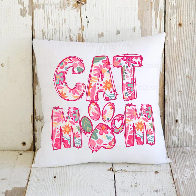 May include: White pillow with the words "CAT MOM" in pink letters outlined in red. The letters are filled with a floral pattern in pink, orange, and green. A pink paw print with a heart in the center is between the words.