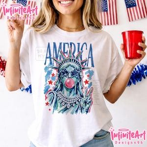 May include: A white t-shirt featuring a graphic illustration of the Statue of Liberty wearing star-shaped sunglasses and blowing a pink bubble. The design includes the word AMERICA in blue, red stars, and a jeweled necklace, rendered in a painterly style.