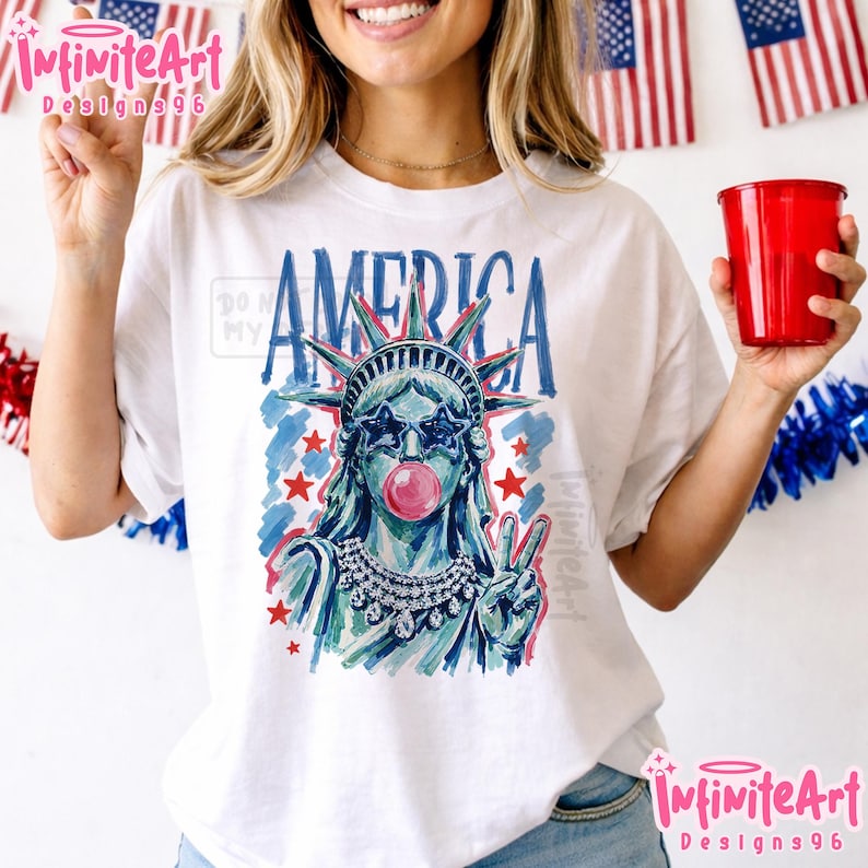 May include: A white t-shirt featuring a graphic illustration of the Statue of Liberty wearing star-shaped sunglasses and blowing a pink bubble. The design includes the word AMERICA in blue, red stars, and a jeweled necklace, rendered in a painterly style.