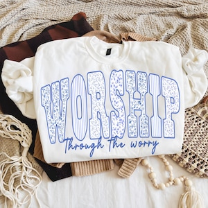 Puede incluir: Sudadera blanca con la palabra "WORSHIP" en letras azules grandes con un estampado floral y rayas verticales. La frase "Through The Worry" está debajo. La sudadera está sobre una superficie blanca con otras prendas.