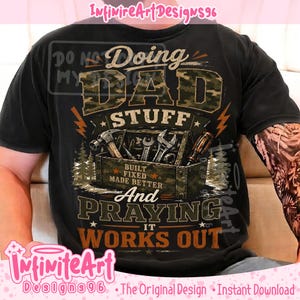 May include: A black t-shirt featuring a graphic with camouflage-patterned text that reads Doing DAD STUFF, a toolbox filled with tools, and the text BUILT FIXED MADE BETTER And PRAYING IT WORKS OUT, accented with orange lightning bolts and pine trees.