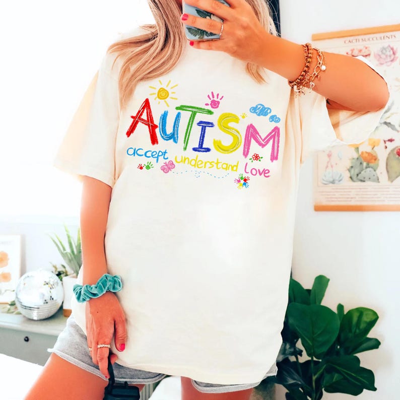 Puede incluir: Camiseta blanca con un gr&aacute;fico colorido que dice "AUTISMO aceptar comprender amar" en una fuente infantil. El gr&aacute;fico incluye un sol, nubes y huellas de manos.