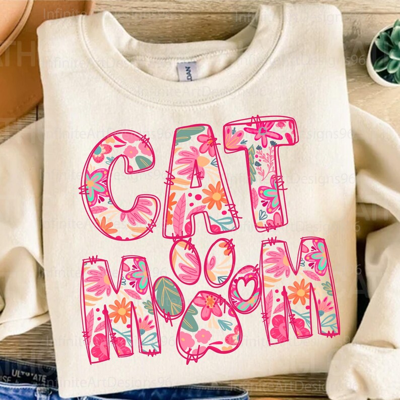 May include: A white sweatshirt with the text "CAT MOM" in pink letters. The letters are decorated with a floral pattern in shades of pink, orange, blue, and green. A pink paw print with a heart in the center is between the words "CAT" and "MOM".