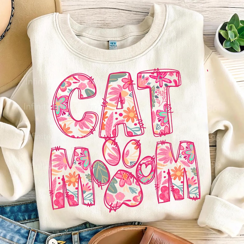 May include: A cream-colored sweatshirt with the words "CAT MOM" in pink outlined letters. The letters are filled with a floral pattern in shades of pink, orange, and green. A pink paw print with a heart in the center is between the words.