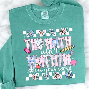 May include: Teal sweatshirt with the text "The Math ain't Mathin' show your work" in a colorful, playful design. Features a checkerboard pattern, flowers, a pencil, an eraser, an apple, and a book.
