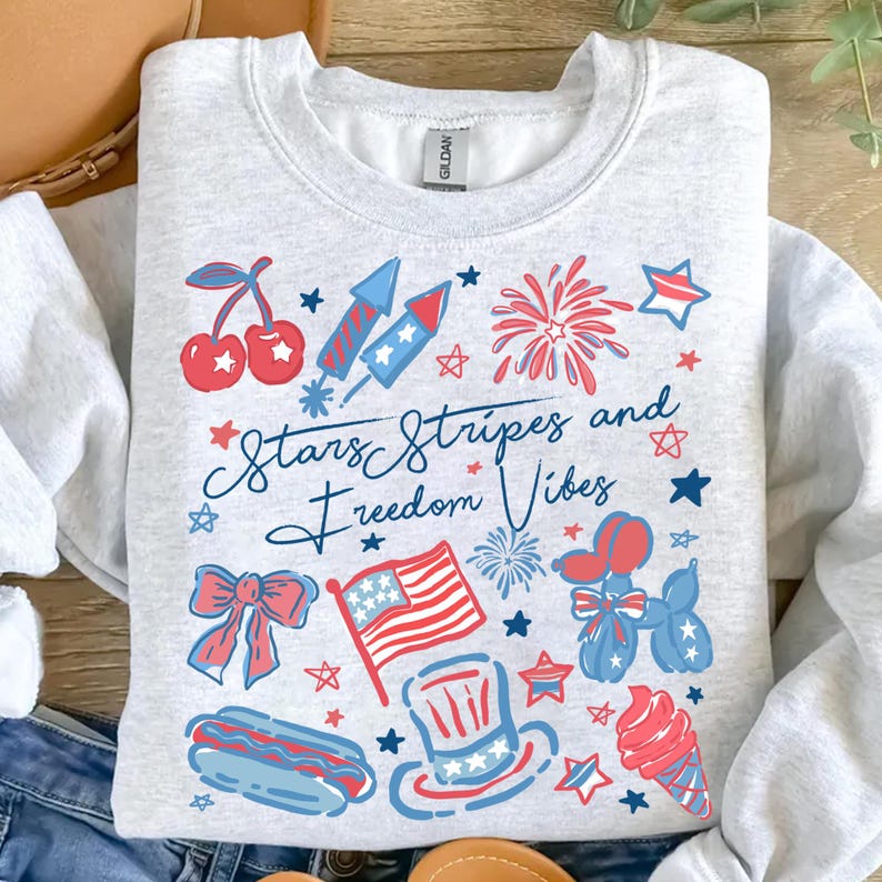 May include: A light gray sweatshirt with a patriotic design. The design includes cherries, fireworks, stars, an American flag, a hot dog, and an ice cream cone. The text "Stars Stripes and Freedom Vibes" is also featured.