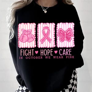 May include: Black sweatshirt with pink graphics of boxing gloves, a ribbon, and a butterfly. The text reads "FIGHT HOPE CARE" and "IN OCTOBER WE WEAR PINK". The design is on a striped background.