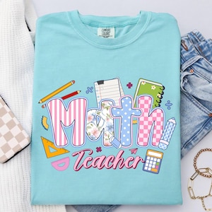 May include: A light blue Comfort Colors t-shirt with a colorful graphic that says "Math Teacher." The word "Math" is in a mix of patterns, including stripes, floral, and checkered designs. The graphic includes pencils, a calculator, and a notebook.