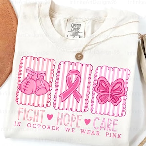 May include: Cream-colored t-shirt with pink graphics and text. The design includes boxing gloves, a ribbon, and a butterfly, all within pink frames. The text reads "FIGHT HOPE CARE" and "IN OCTOBER WE WEAR PINK".