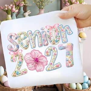 Puede incluir: Una tela blanca con la inscripci&oacute;n bordada "SENIOR 2021". Las letras y los n&uacute;meros est&aacute;n decorados con motivos florales, patchwork y lazos. Una gran flor rosa est&aacute; centrada debajo de la palabra "SENIOR".