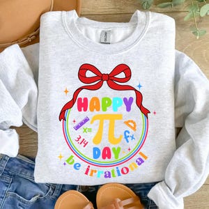 May include: A gray sweatshirt with a colorful graphic design that says "Happy Pi Day be irrational". The graphic features a red bow, a rainbow circle, and math symbols.