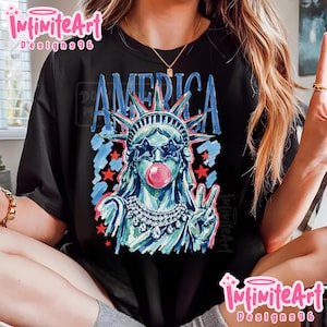 May include: A black t-shirt featuring a graphic of the Statue of Liberty wearing star-shaped sunglasses and blowing a pink bubble. The design includes the word AMERICA in blue, red stars, and a jeweled necklace, rendered in a painterly style.
