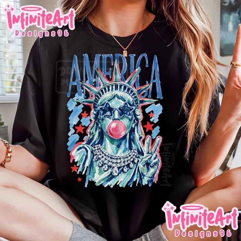 May include: A black t-shirt featuring a graphic of the Statue of Liberty wearing star-shaped sunglasses and blowing a pink bubble. The design includes the word AMERICA in blue, red stars, and a jeweled necklace, rendered in a painterly style.