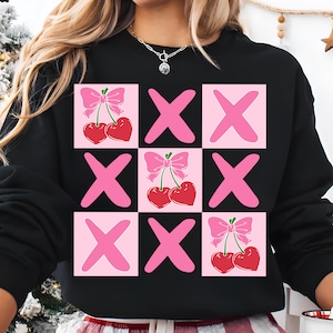 Könnte beinhalten: Schwarzes Sweatshirt mit einem rosa und roten Grafikdesign. Das Design zeigt rosa X's, rosa Quadrate und rote Kirschen mit rosa Schleifen. Ein lässiges Kleidungsstück für jeden Tag.