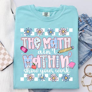 May include: A light blue t-shirt with the text "The Math ain't nothin' show your work" in a colorful, playful font. The design includes flowers, a pencil, an eraser, an apple, and a book. The shirt is folded on a white surface.