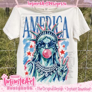 May include: A cream-coloured short-sleeve t-shirt featuring a graphic of the Statue of Liberty wearing star-shaped sunglasses and blowing a pink bubble. The design includes the word AMERICA in blue, red stars, and a jeweled necklace illustration.