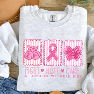May include: Light gray sweatshirt with pink graphics. The design includes boxing gloves, a ribbon, and a butterfly, all within pink striped frames. Text reads "FIGHT HOPE CARE" and "IN OCTOBER WE WEAR PINK".