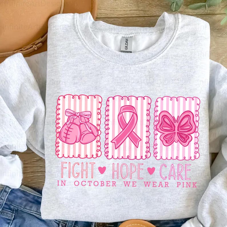 May include: Light gray sweatshirt with pink graphics. The design includes boxing gloves, a ribbon, and a butterfly, all within pink striped frames. Text reads "FIGHT HOPE CARE" and "IN OCTOBER WE WEAR PINK".