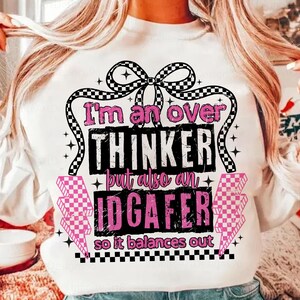 May include: White sweatshirt with the text "I'm an over thinker but also an IDGAFer so it balances out" in pink and black. The design includes a checkered bow, lightning bolts, and stars.